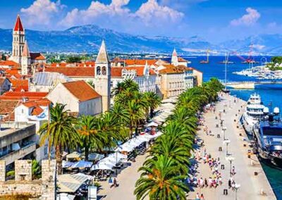 Promenade in Trogir, Croatia, with palm trees, historic buildings, and vibrant seaside activity.