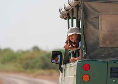 A young child wearing a safari hat leaning out of a green safari vehicle, flashing a peace sign, representing the joy and family-friendly nature of wildlife safaris.